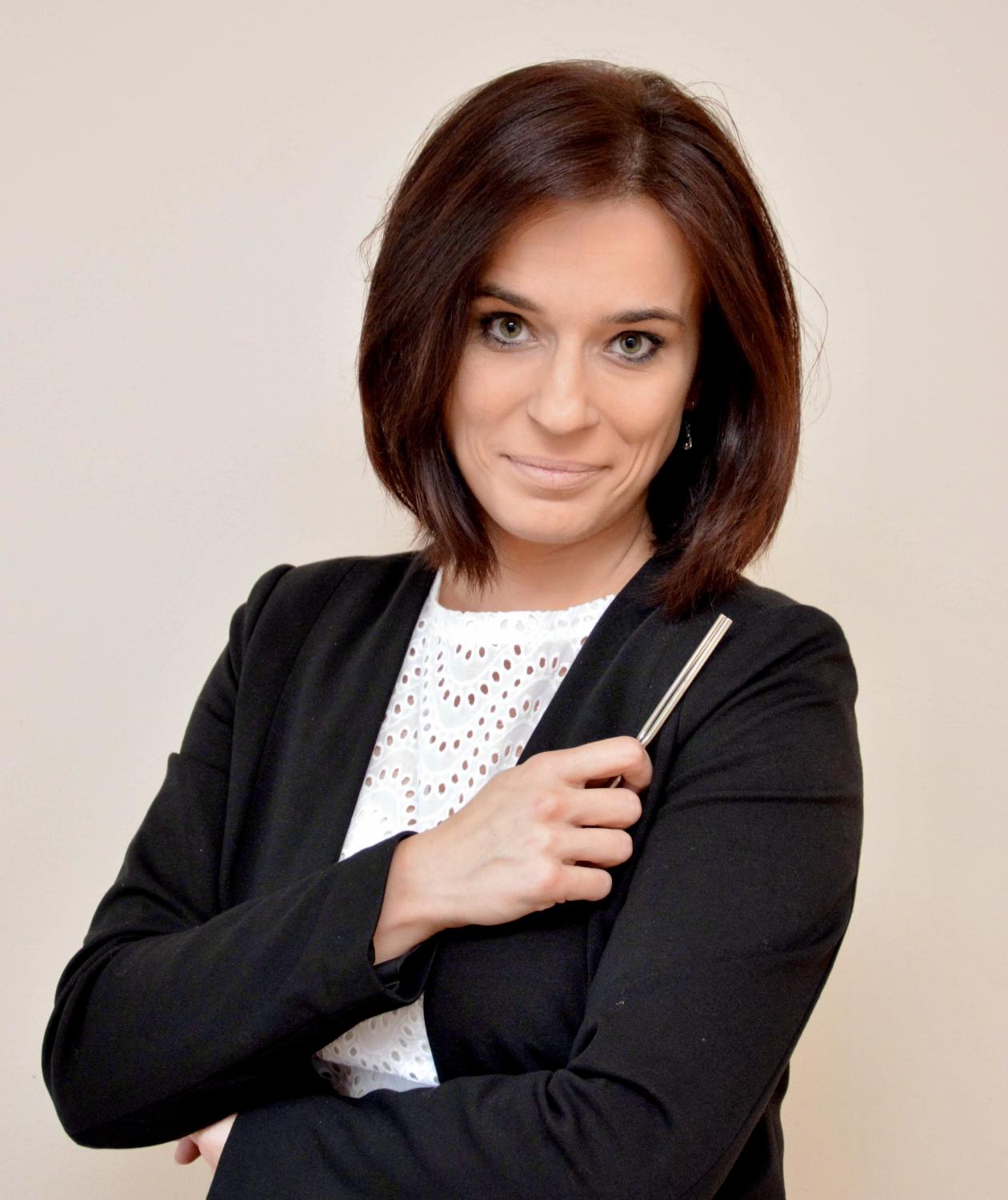 Dyryentka chóru trzyma w lewej ręce batutę, jest ubrana w czarny żakiet i białą bluzkę