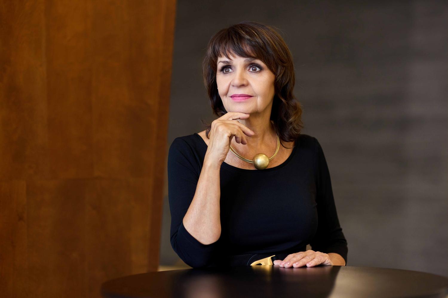 Dyrygentka Anna Szotka ubrana jest w czarną sukienkę, nosi czrne włosy i wyciąga lewą rękę przed siebie