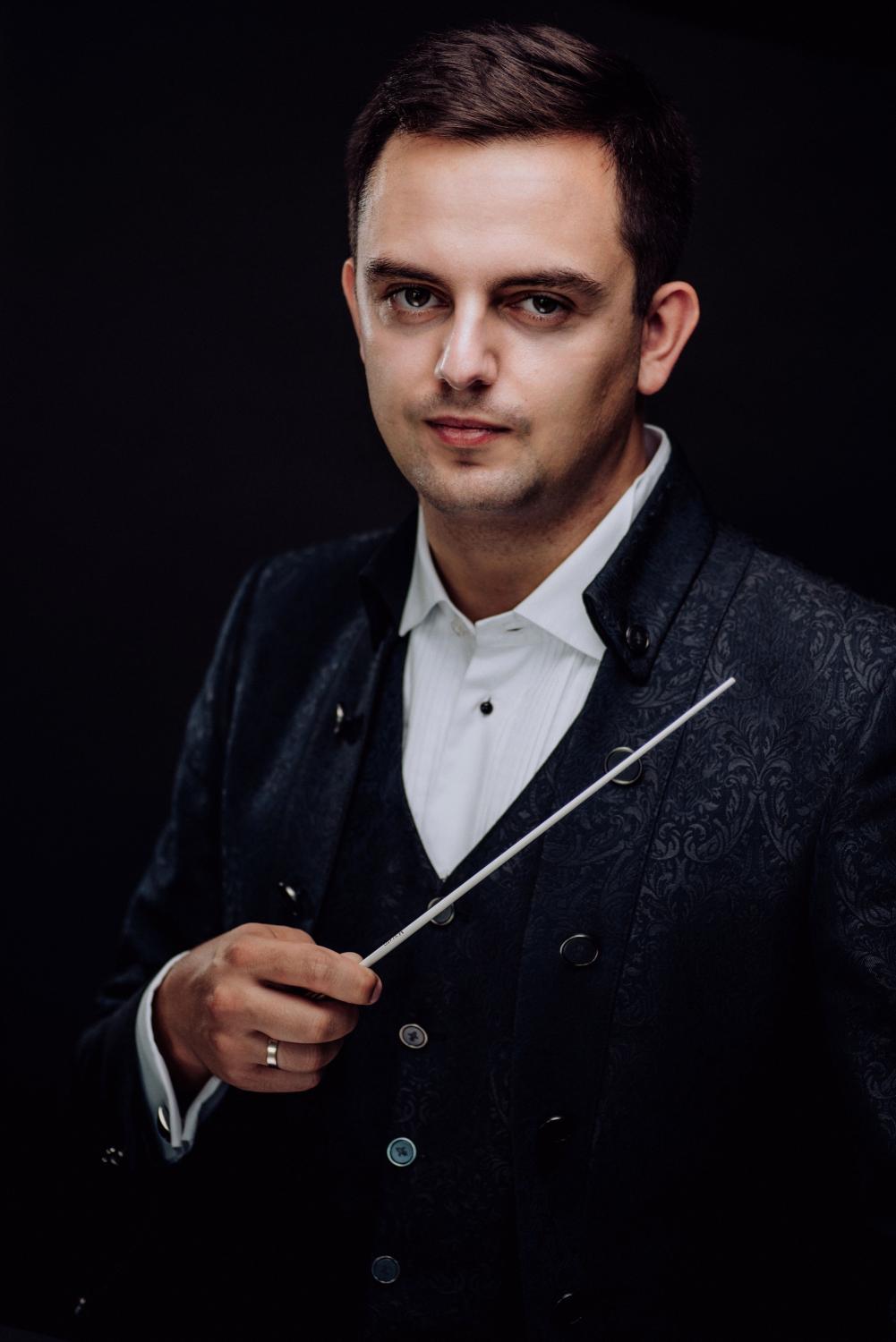 Młody dyrygent Maciej Tomasiewicz trzyma w oprawej ręce batutę i ubrany jest w szarą ozdobną marybarkę i białą koszulę