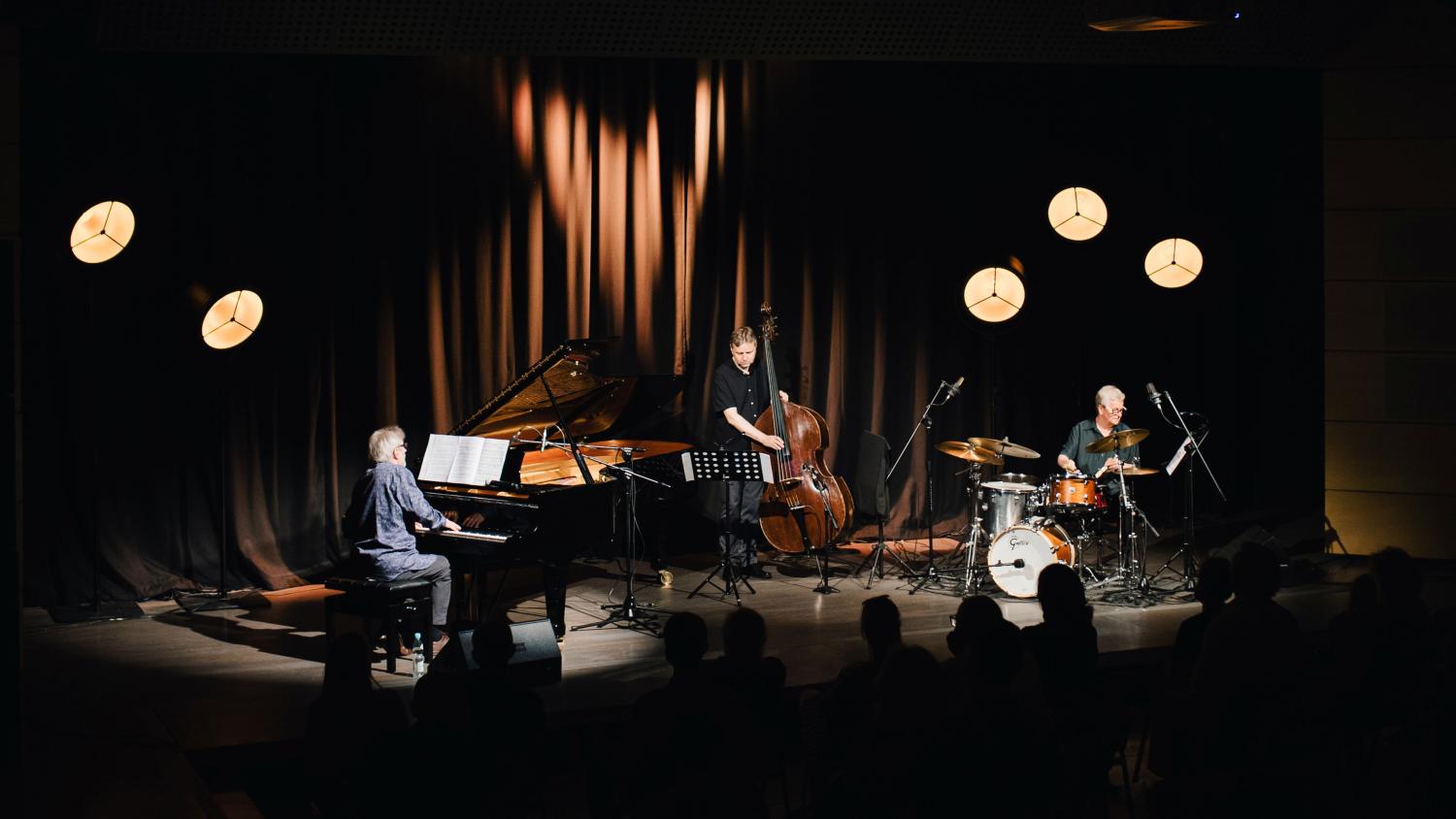 Trio Andrzeja Jagodzińskiego, pianista siedzi przy czarnym fortepianie, kontrabasista stoi z instrumentem, perkusista siedzi za składem instrumentów