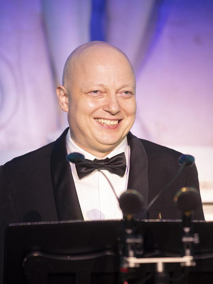 Śpiewak Witold Żołądkiewicz ubrany jest w czarny smoking i czarną muchę, ma łysinę, stoi uśmiechnięty przed mikrofonemm